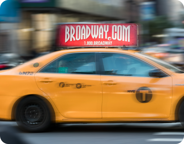 A cab in New York City drives down the street with the Broadway.com logo advertisement on top